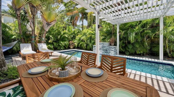 Eine Holzterrasse mit Pool, umgeben von üppigen tropischen Pflanzen und einer weißen Pergola.