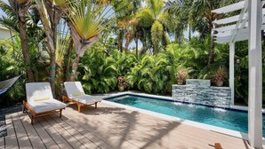 Un área de piscina con dos sillas de descanso, rodeada de exuberante vegetación tropical y una pérgola blanca.