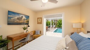 Ein Schlafzimmer mit Bett, Schreibtisch, Stuhl und Fernseher. Eine Tür führt zu einem Poolbereich mit Gartenmöbeln.