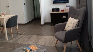 Un soggiorno moderno con una poltrona grigia, un tavolino rotondo con una rivista, una zona pranzo con tavolo e sedie, una TV a schermo piatto montata a parete e una porta che dà su un'altra stanza.