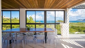 A dining area with a wooden ceiling, large windows with a view of the sea, and clear glass chairs. - La Siesta (4/5 Rooms) (La Baie Nettle)