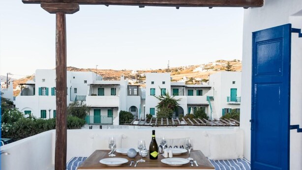A terrace with a table set for two, overlooking a row of white buildings with green shutters. - Glamorous 2BR Villa in Ornos w Sea View (Mykonos)