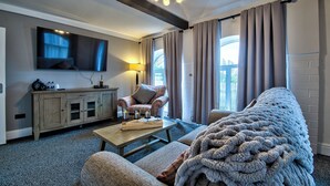 A living room with a sofa, a coffee table, a TV mounted on the wall, a fireplace, and a window with curtains. - The George Of Piercebridge (Darlington)