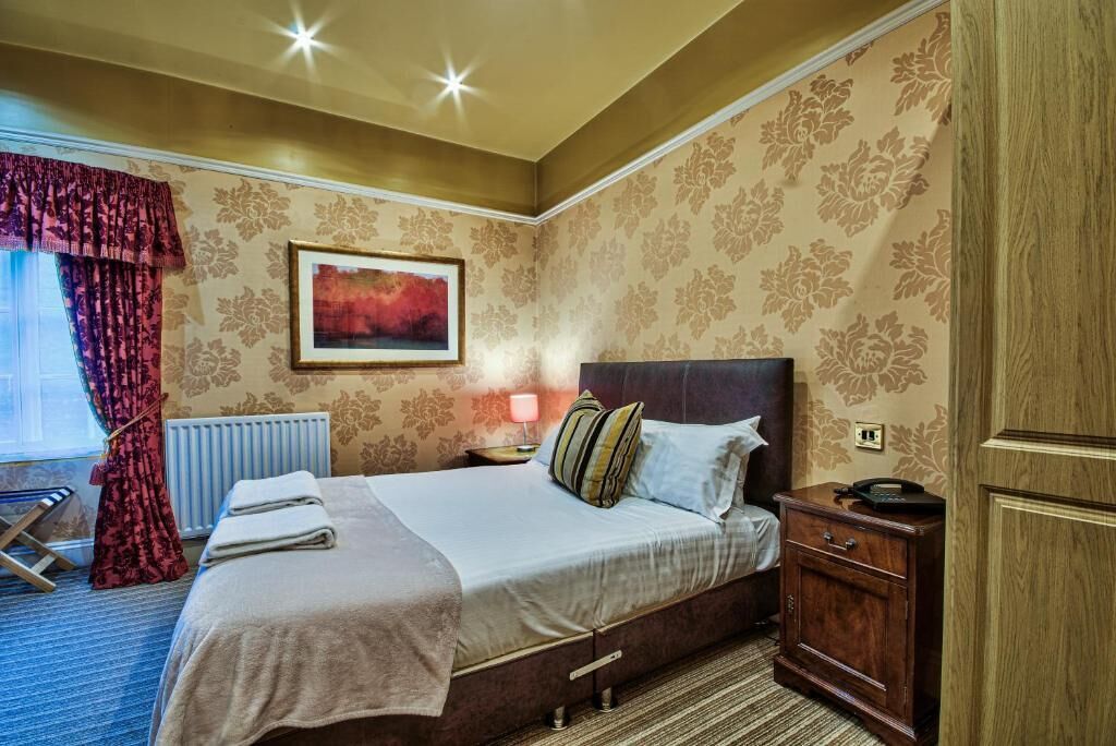 A hotel room with a bed, a nightstand, a lamp, a radiator, and a window with curtains.