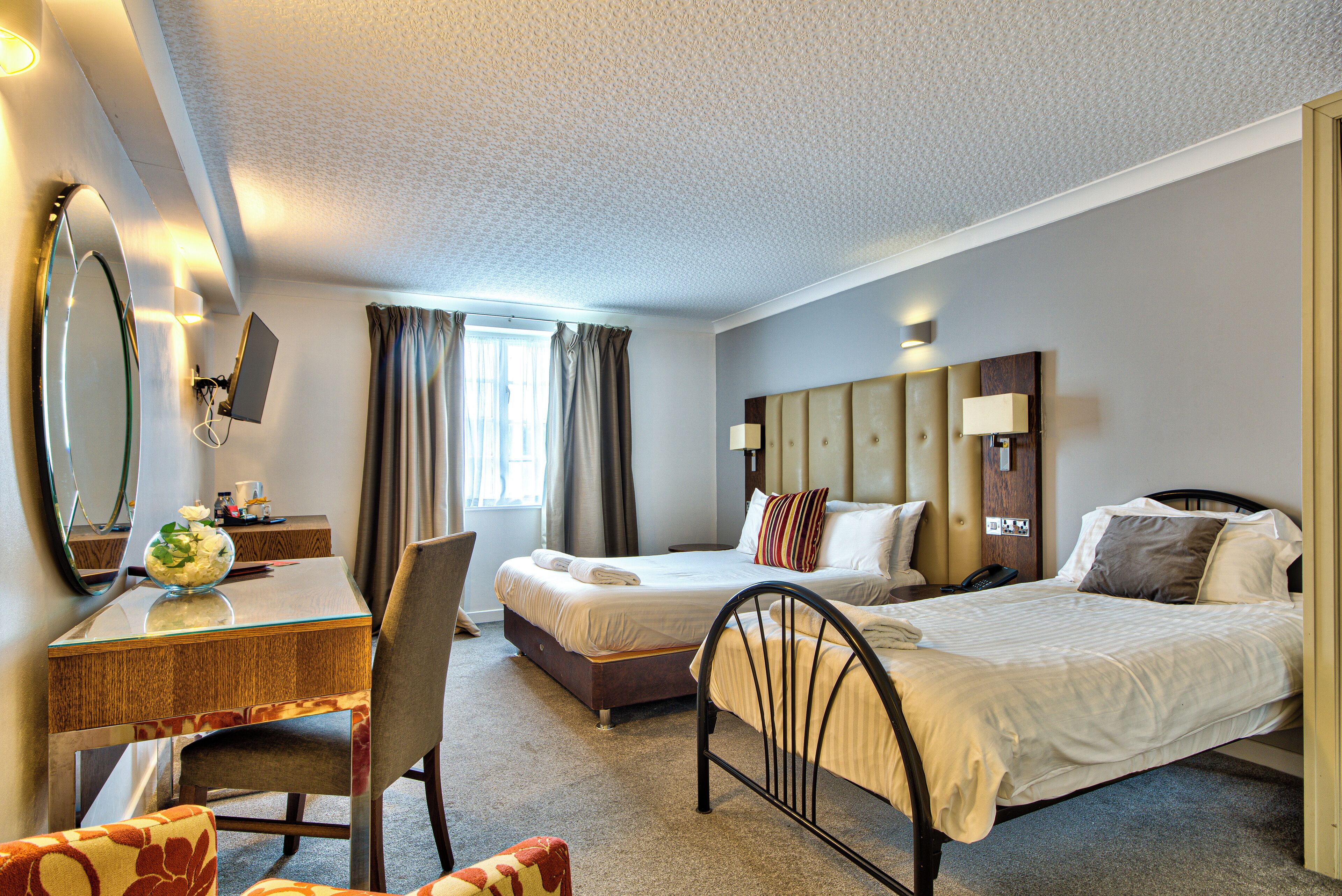 A hotel room with a bed, a desk with a chair, a mirror, and a window with curtains.