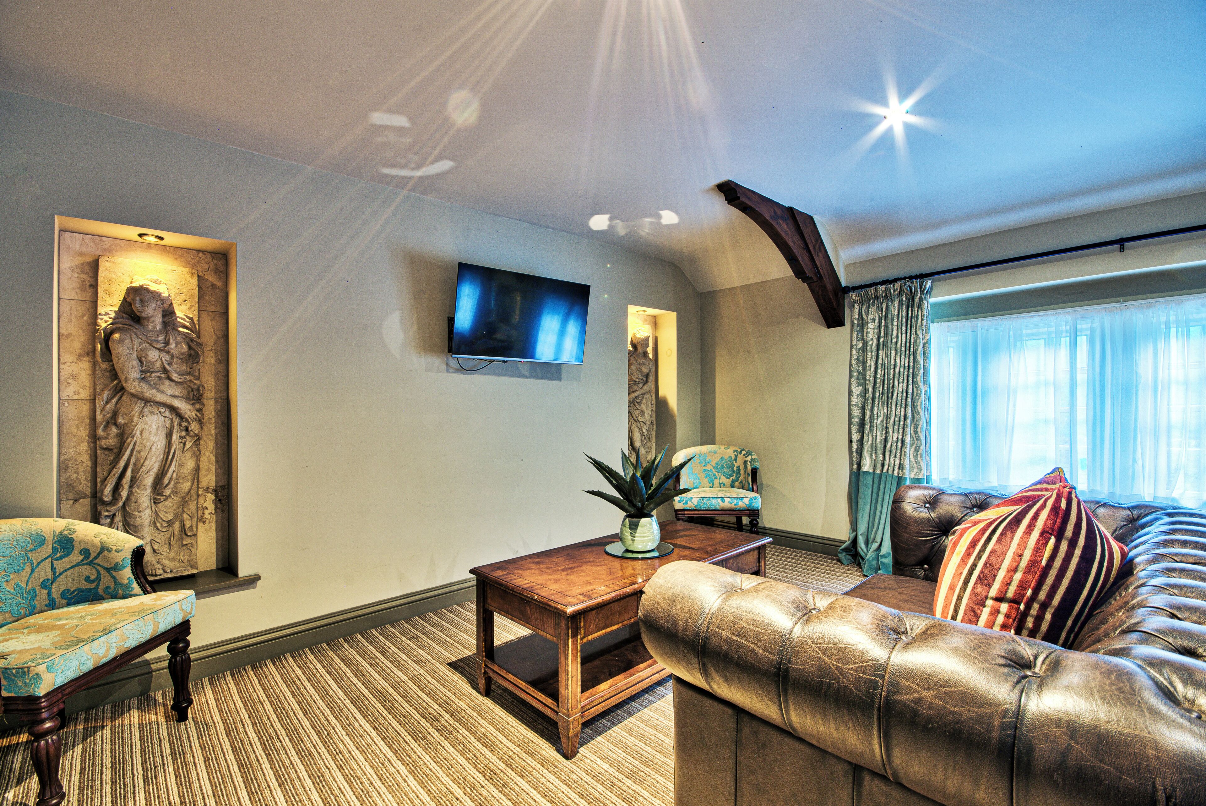 A living room with a sofa, armchair, a flat-screen TV, a statue, and a window with curtains.