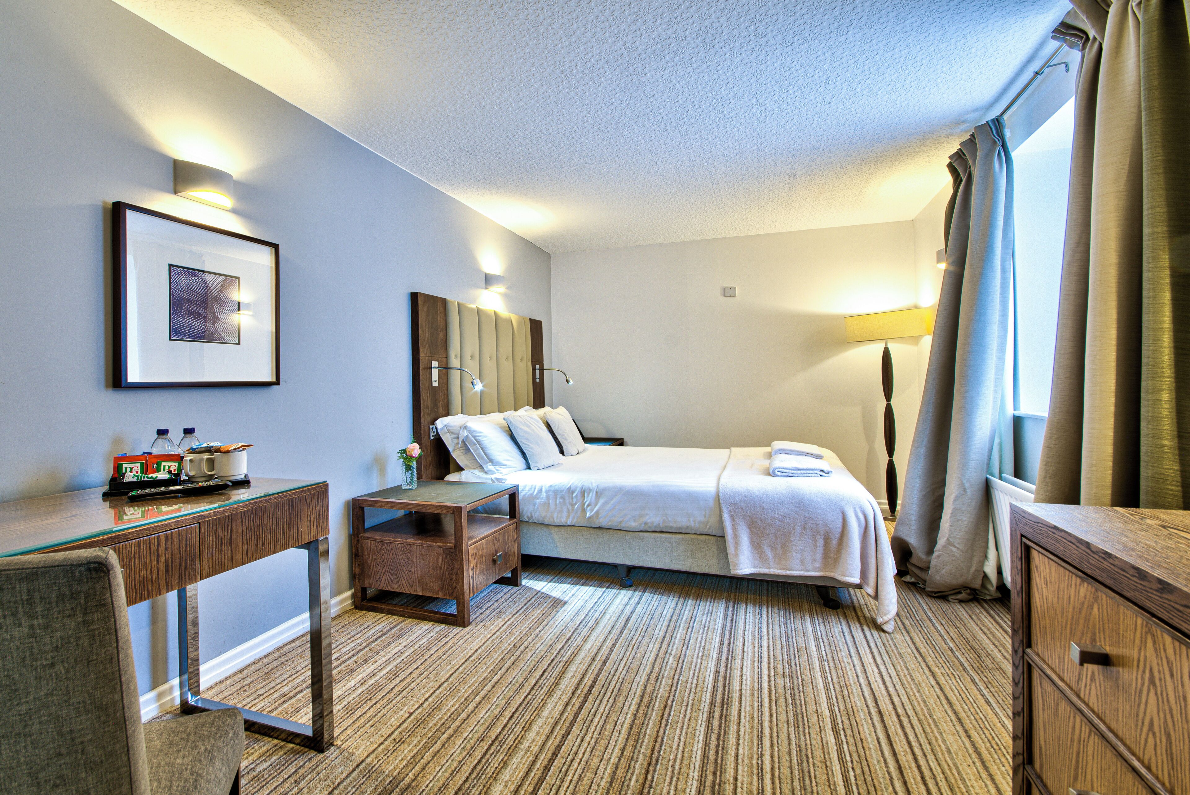 Ein Hotelzimmer mit Bett, Nachttisch, Lampe und einem gerahmten Bild an der Wand.