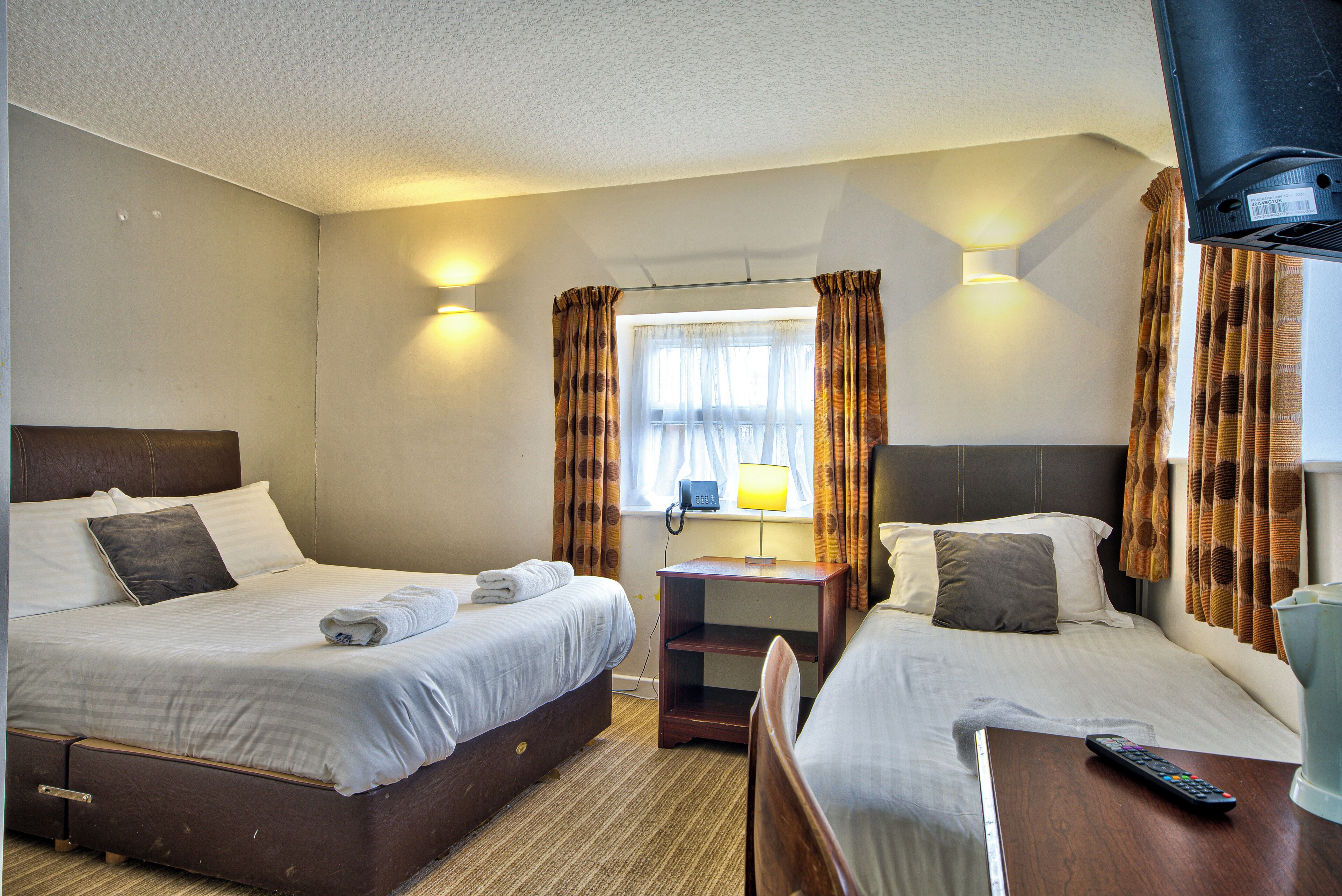 Ein Hotelzimmer mit zwei Betten, einem Nachttisch, einer Lampe, einer Fernbedienung und einem Fenster mit Vorhängen.