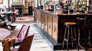 A bar area with wooden bar stools, a counter with a cash register, and a variety of bottles on shelves. - The George Of Piercebridge (Darlington)