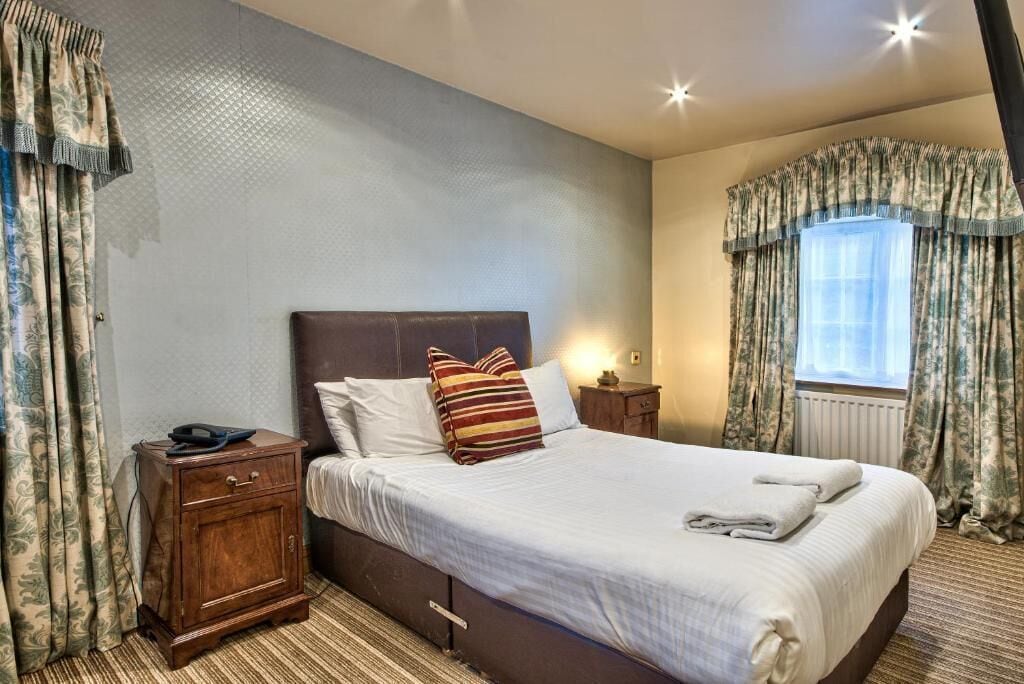 A neatly made bed with pillows, a bedside table with a lamp, and a window with curtains.