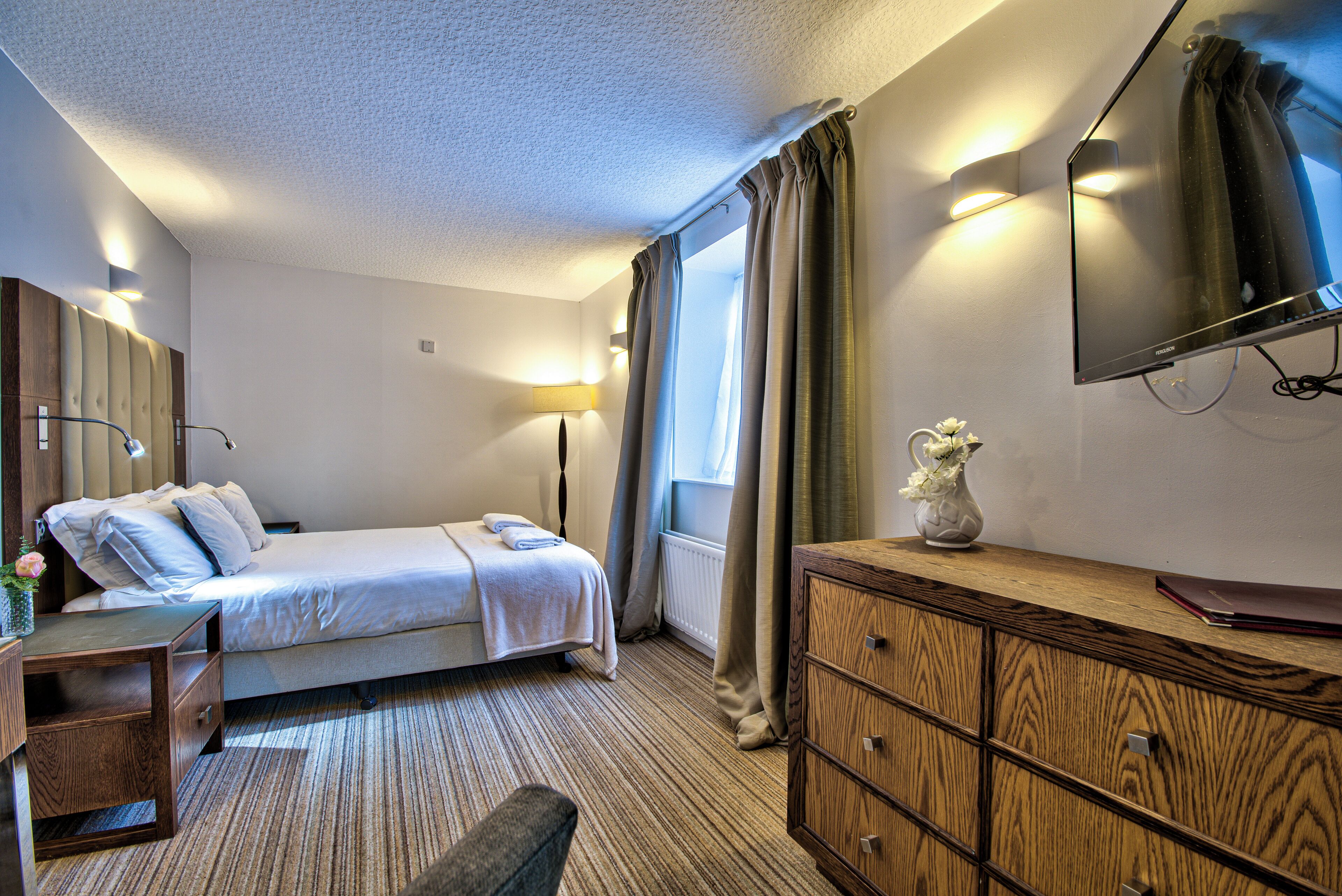 A hotel room with a bed, bedside table, lamp, dresser, and a flat-screen TV mounted on the wall.