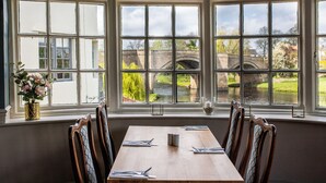 A dining area with a table set for four, large windows with a view of a bridge and greenery, and a vase with flowers on the windowsill. - The George Of Piercebridge (Darlington)