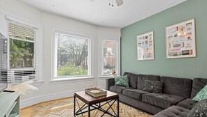 Ein Wohnzimmer mit grauem Sofa, einem Holztisch und einem Teppich mit Zickzackmuster. Drei Fenster mit weißen Jalousien und zwei gerahmte Bilder sind an der Wand zu sehen.