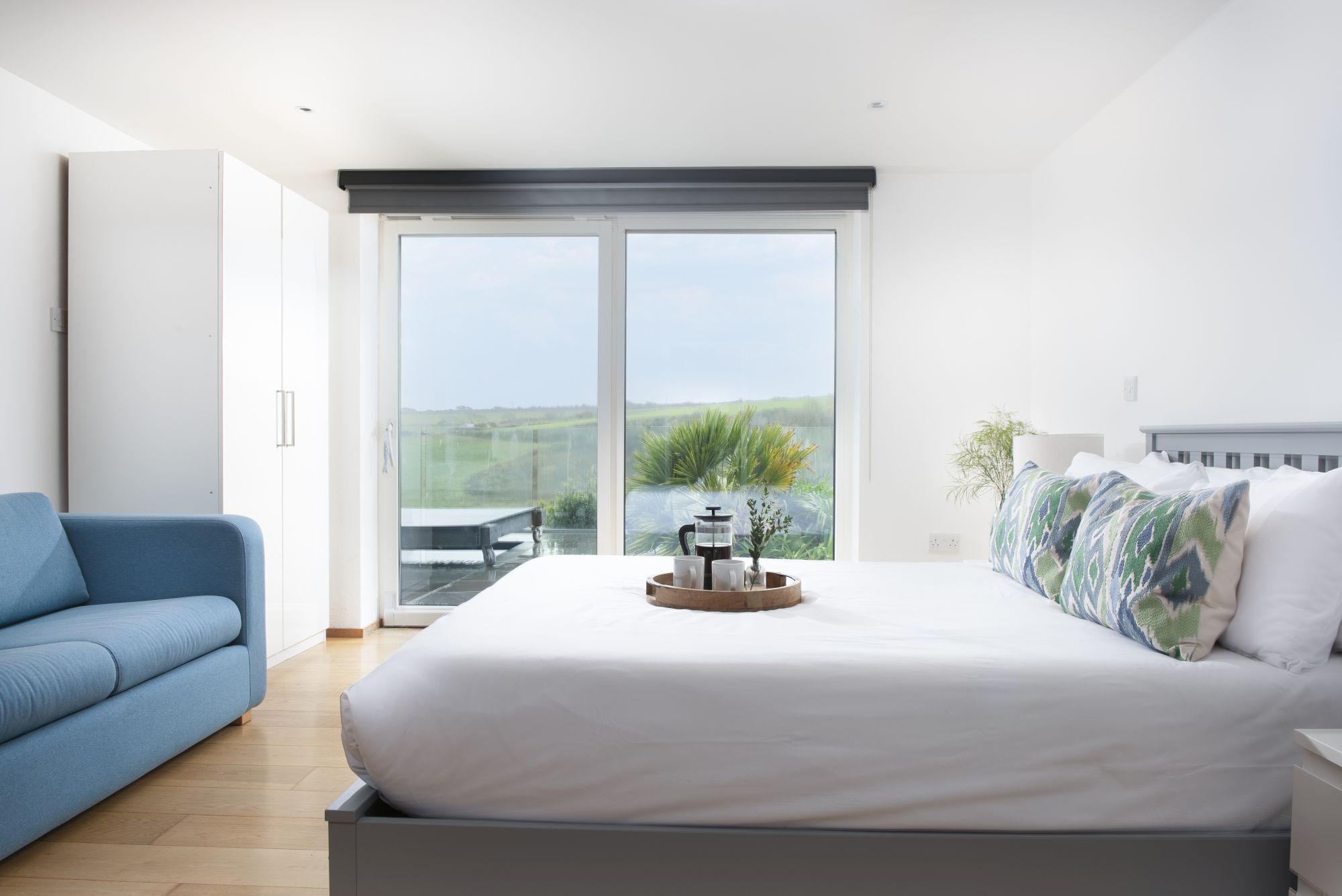 A bedroom with a large bed, a blue sofa, a coffee table with a tray, and a view of greenery through a sliding door.