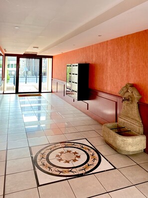 Un vestíbulo amplio con suelo de baldosas, un diseño circular decorativo, una fuente de piedra y una fila de taquillas frente a una pared naranja.