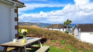 A wooden picnic table with a vase of flowers and a mug, overlooking a coastal view with houses and a clear sky. - C41 Windmills, Riviere Towans (Hayle)