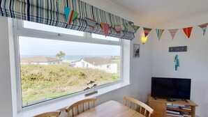 A room with a large window overlooking a beach, a television on a wooden stand, and a small table with chairs. - C41 Windmills, Riviere Towans (Hayle)