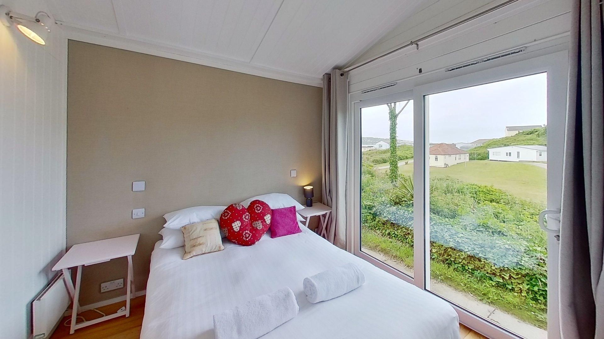 A bedroom with a large window, a bed with white bedding, a bedside table, and a lamp.