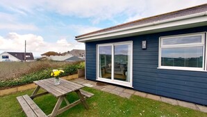 Une maison à l’extérieur d’un bleu foncé, avec une terrasse en bois et une table de patio agrémentée d’un vase de fleurs jaunes.
