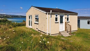 Une petite maison à un seul étage, à l’extérieur blanc et au toit foncé, située sur une colline verdoyante, d’où l’on peut admirer la mer et les collines en arrière-plan.
