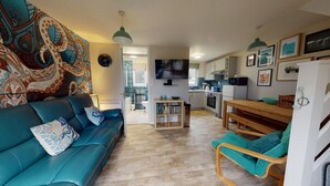 A living room with a blue sofa, a wooden dining table, and a kitchen area with a microwave and stove. - 45 Strawberry Hill, Tolroy Manor (Hayle)