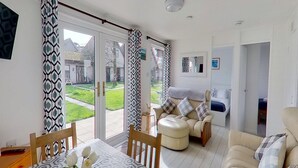 A spacious living area with a sliding glass door leading to a garden, a sofa, a dining table, and a small side table. - 25 Trevithick Court, Tolroy Manor (Hayle)