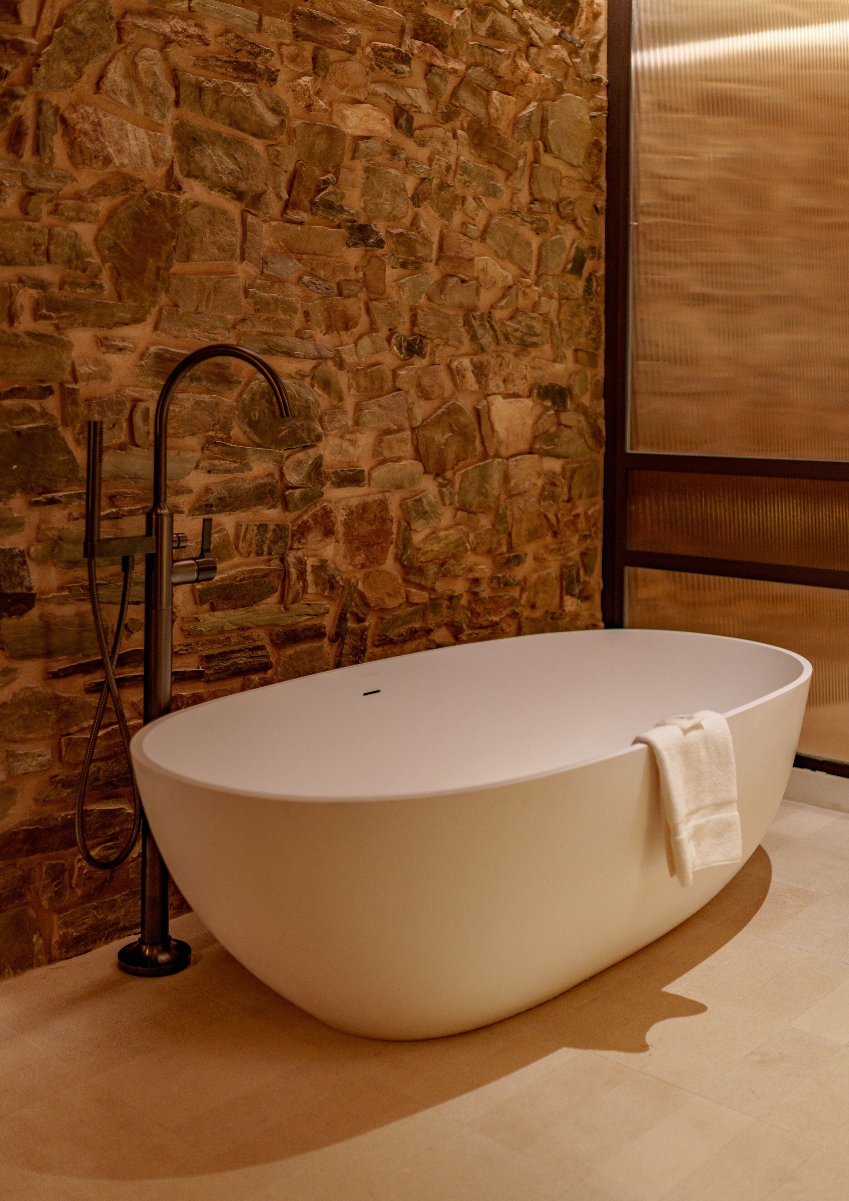 Un baño moderno con una bañera independiente, una pared de piedra y un ventanal amplio.
