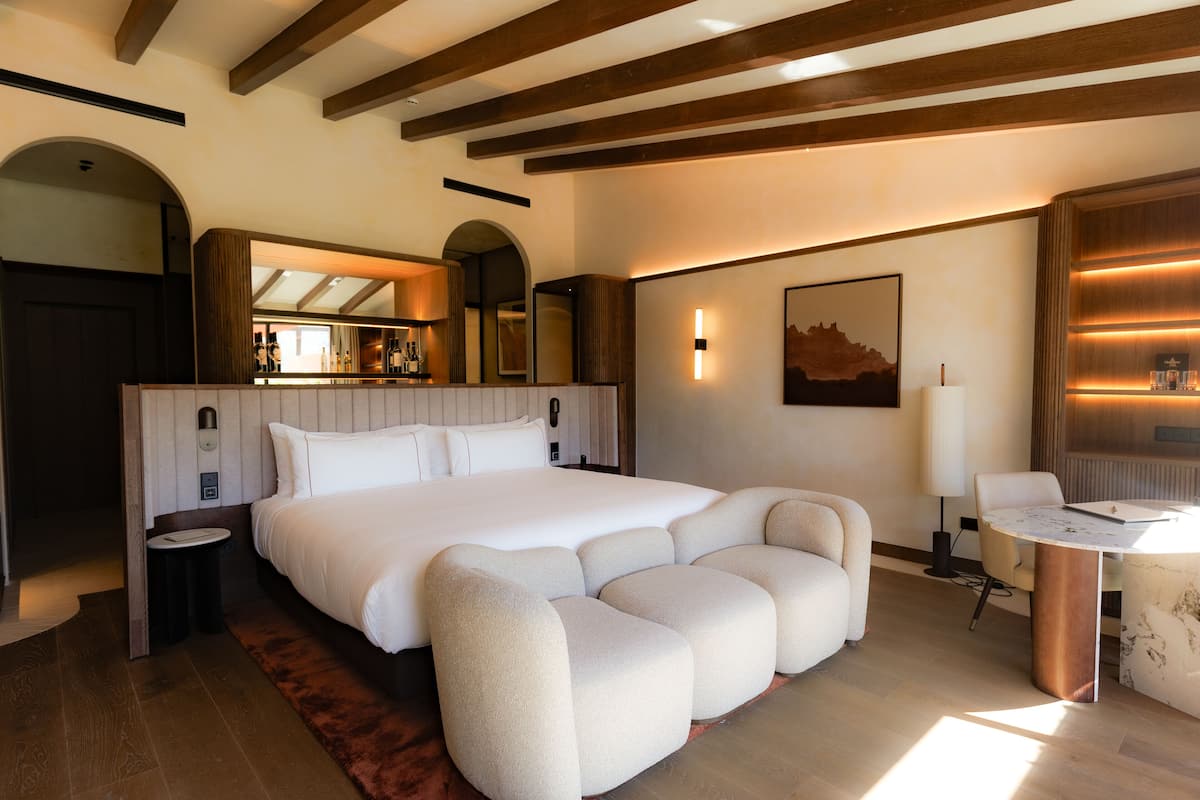 A modern hotel room with a large bed, a seating area, a small table, and a bathroom visible through an archway.