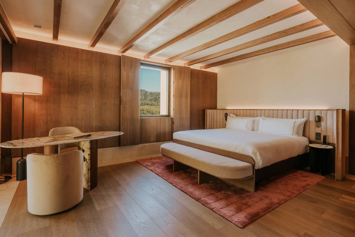 A bedroom with a wooden ceiling, a large bed, a desk, and a window.