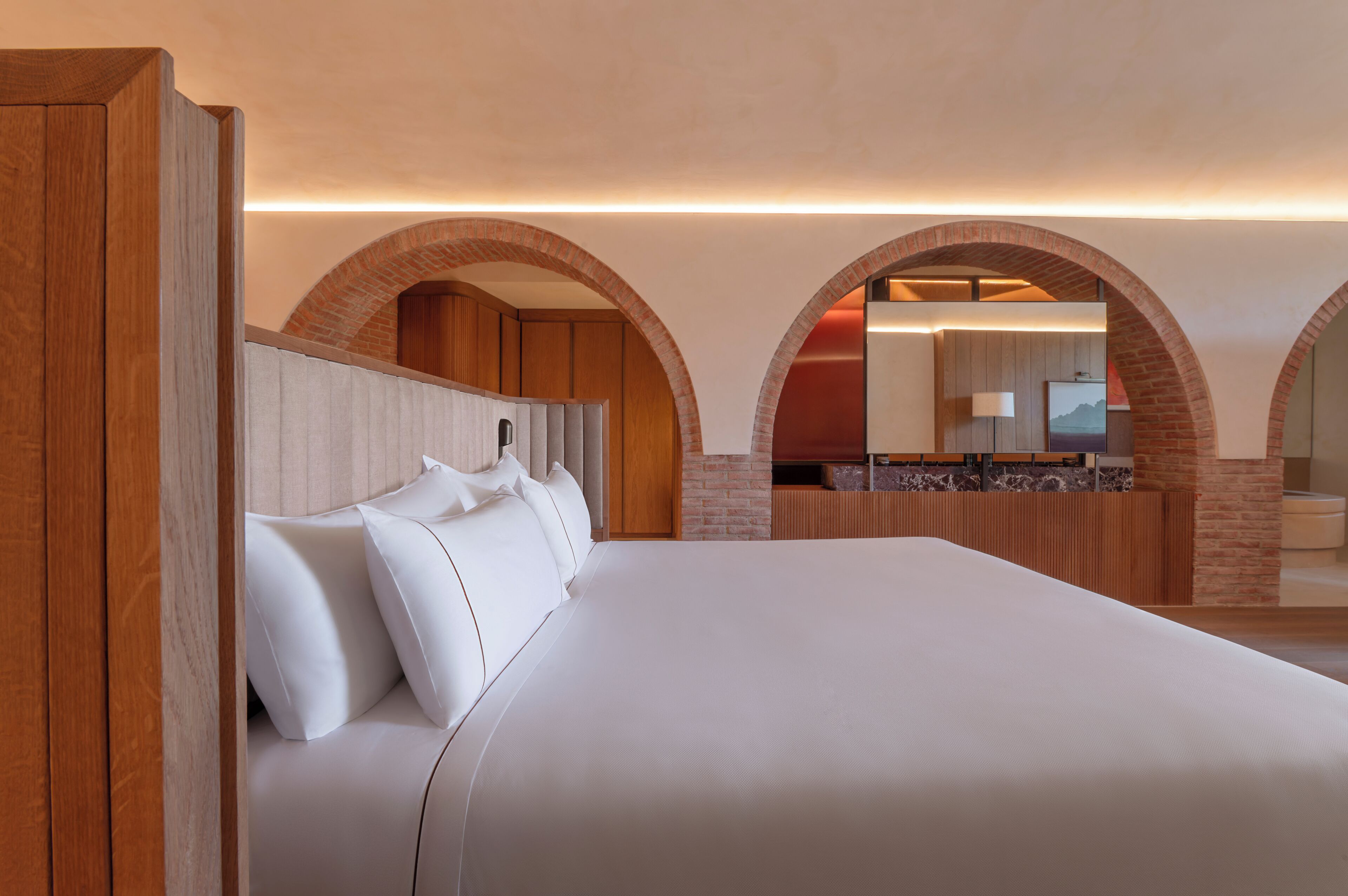 Una cama grande con sábanas blancas, cabecera de madera y un arco de ladrillo que da a un baño.