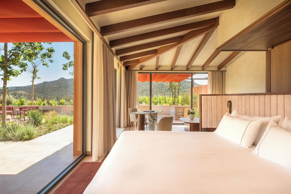 A modern hotel room with a large bed, a dining area, and a view of the outdoors.
