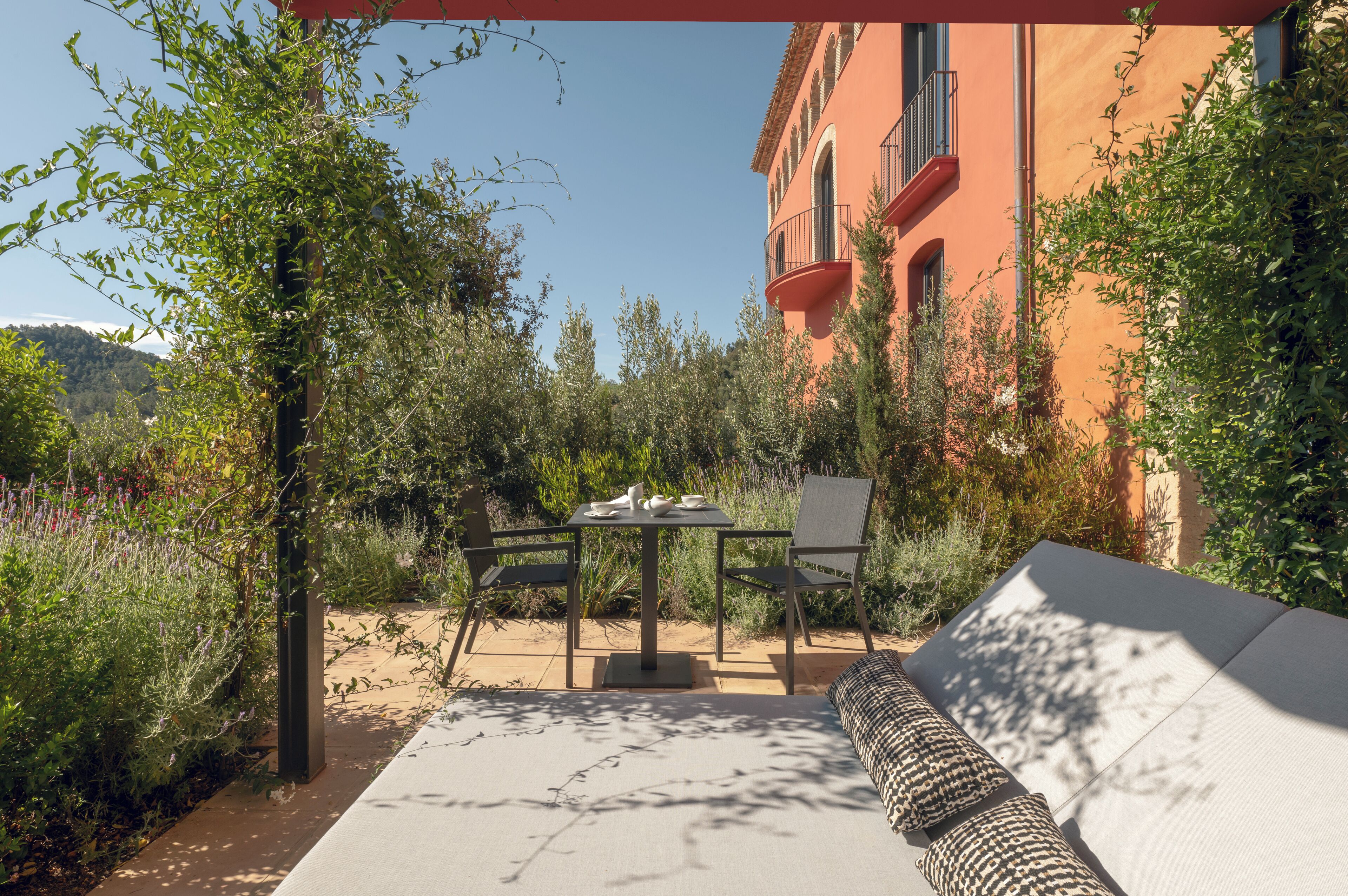 An outdoor patio with a table and chairs, surrounded by greenery and a building with red walls.