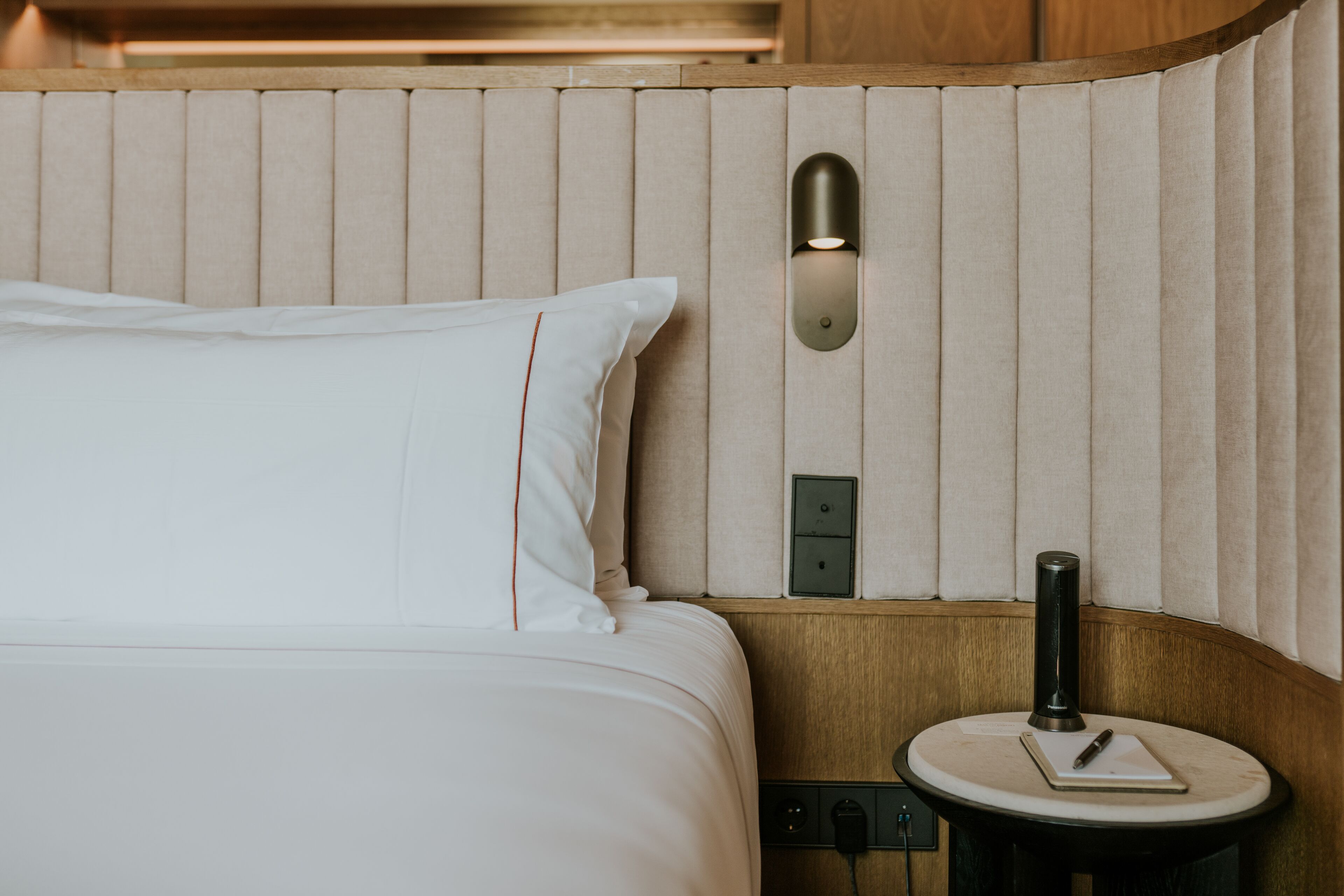A neatly made bed with a pillow, a bedside table with a lamp, and a wall-mounted light fixture.