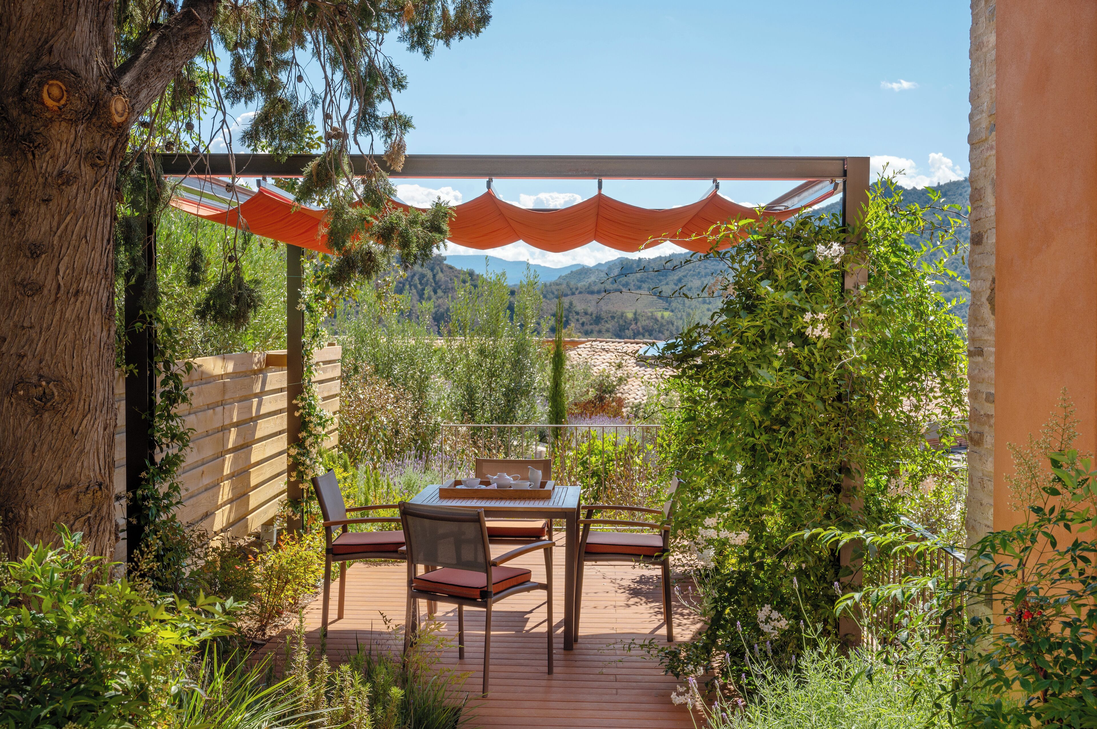 Um deck de madeira com uma mesa e cadeiras, rodeado por vegetação e vista para montanhas.