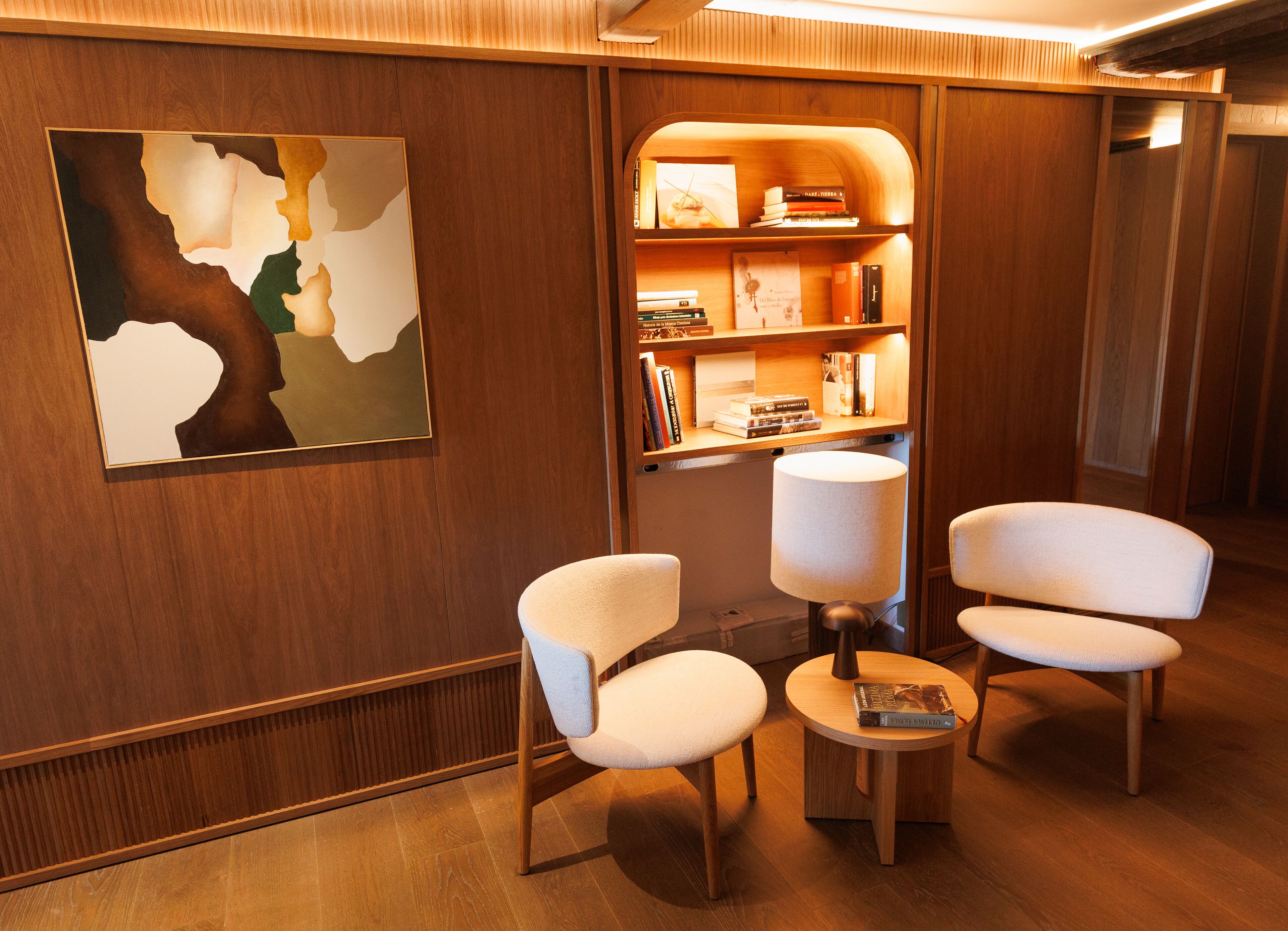 Eine moderne Hotellobby mit Holzvertäfelung, einem großen abstrakten Gemälde, zwei weißen Sesseln, einem kleinen runden Tisch mit einer Lampe und einem Bücherregal mit Büchern und Dekoration.