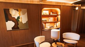 Eine moderne Hotellobby mit Holzvertäfelung, einem großen abstrakten Gemälde, zwei weißen Sesseln, einem kleinen runden Tisch mit einer Lampe und einem Bücherregal mit Büchern und Dekoration.