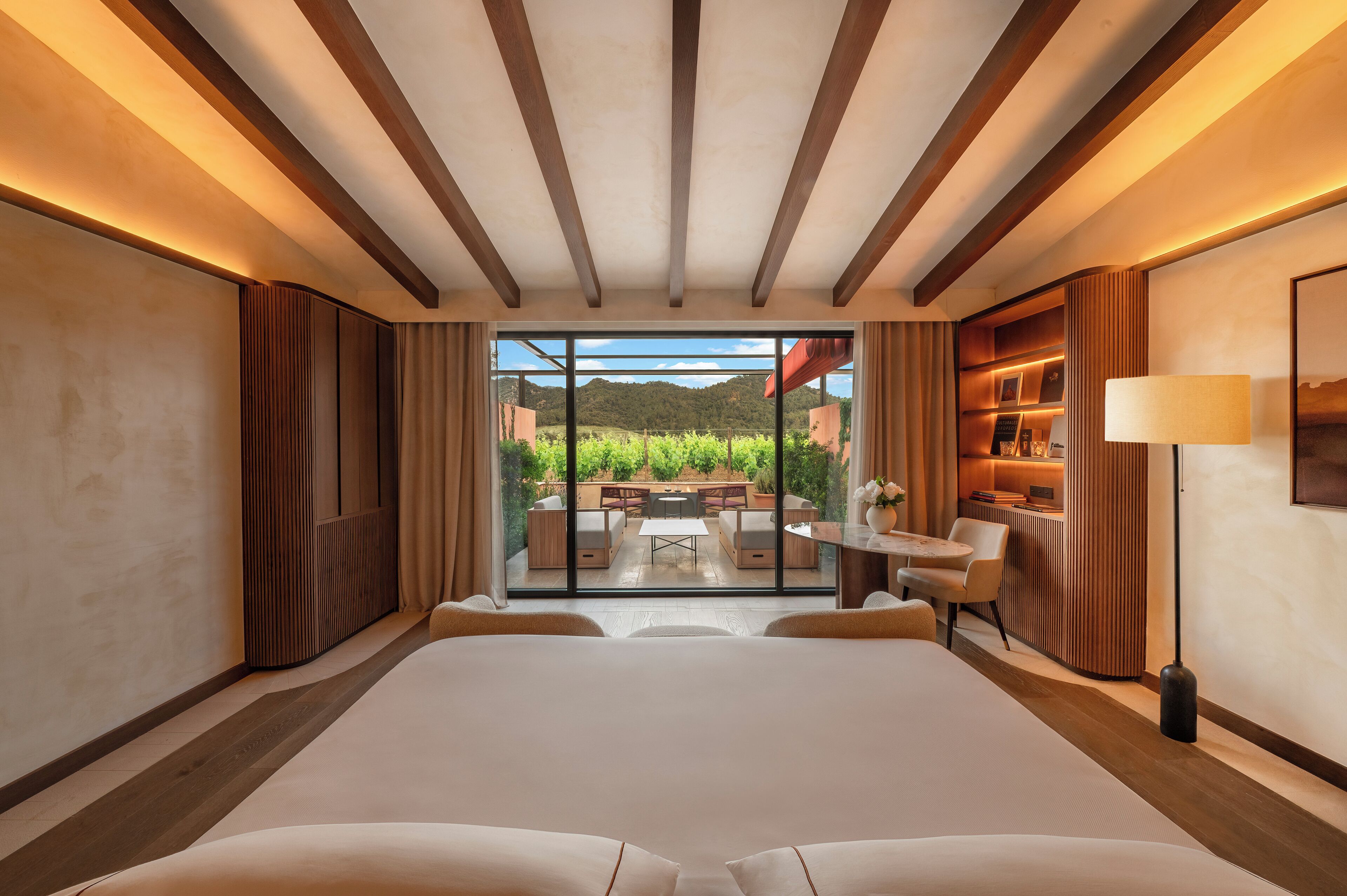 A modern hotel room with a large bed, a view of a garden and mountains, and a cozy seating area with a table and chairs.