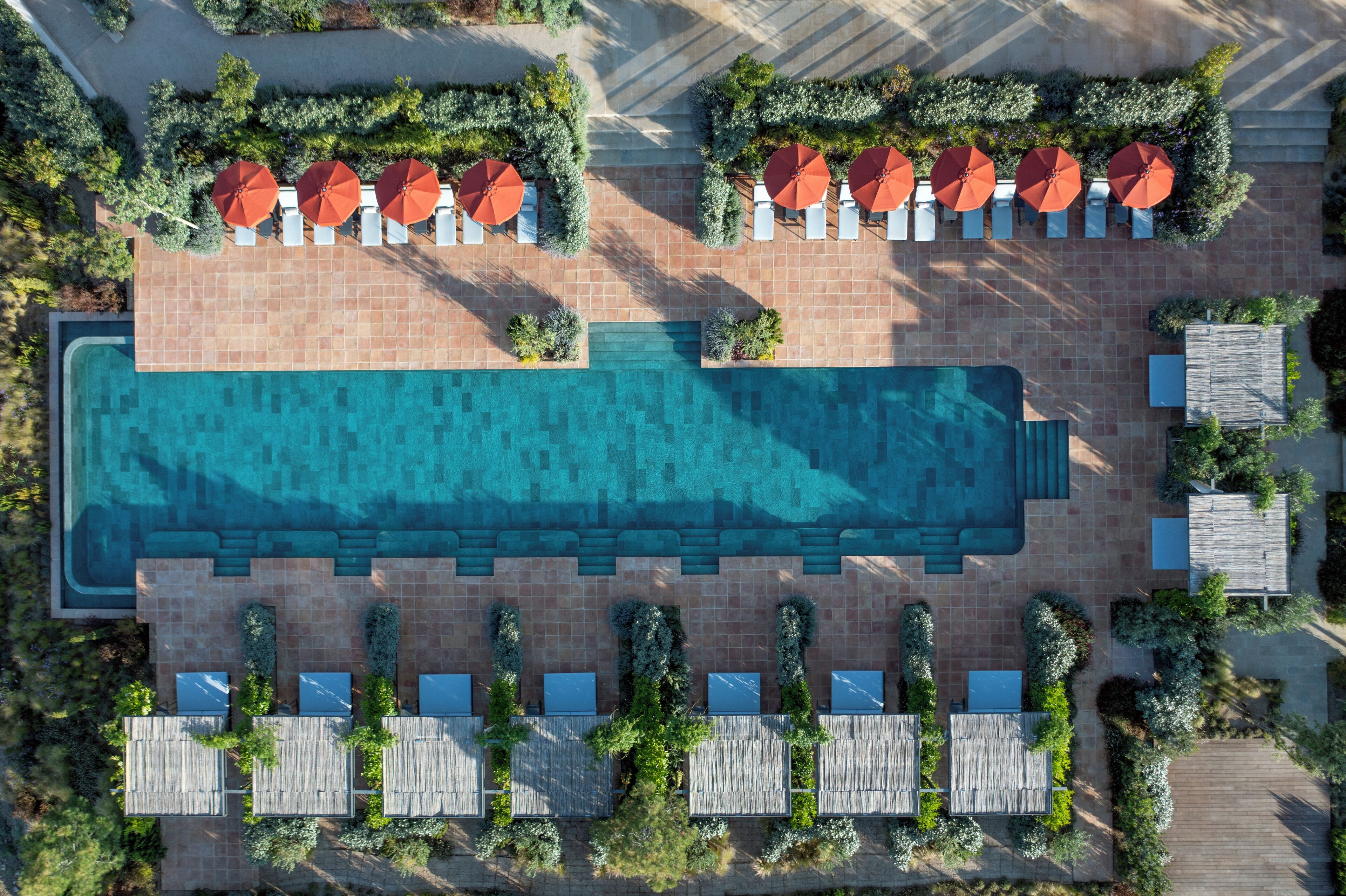 An aerial view of a resort with a large swimming pool, multiple lounge areas with umbrellas, and a well-maintained landscape.