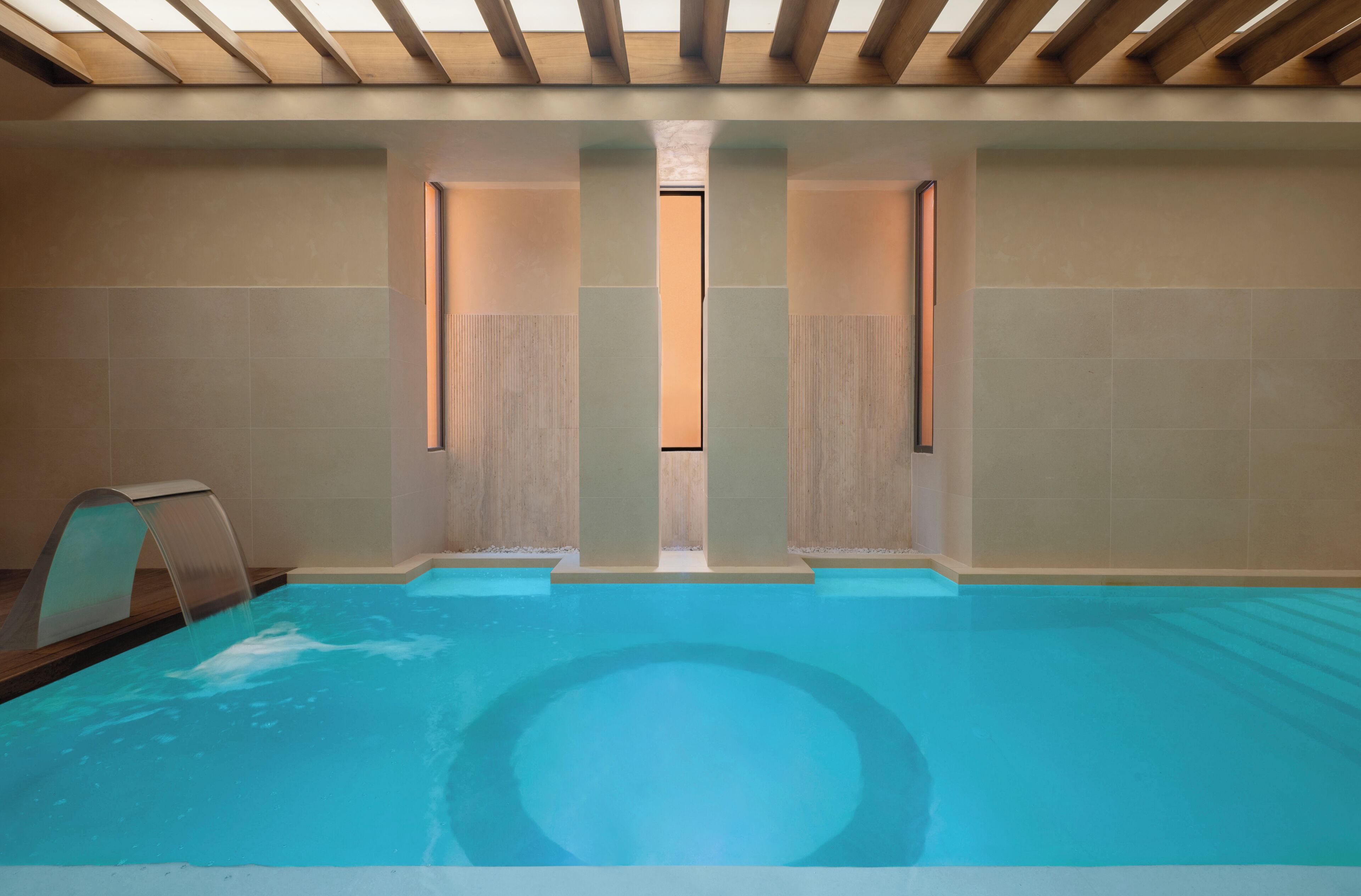 Una piscina interior moderna con una fuente de agua cristalina, vigas de madera en el techo y un diseño minimalista.