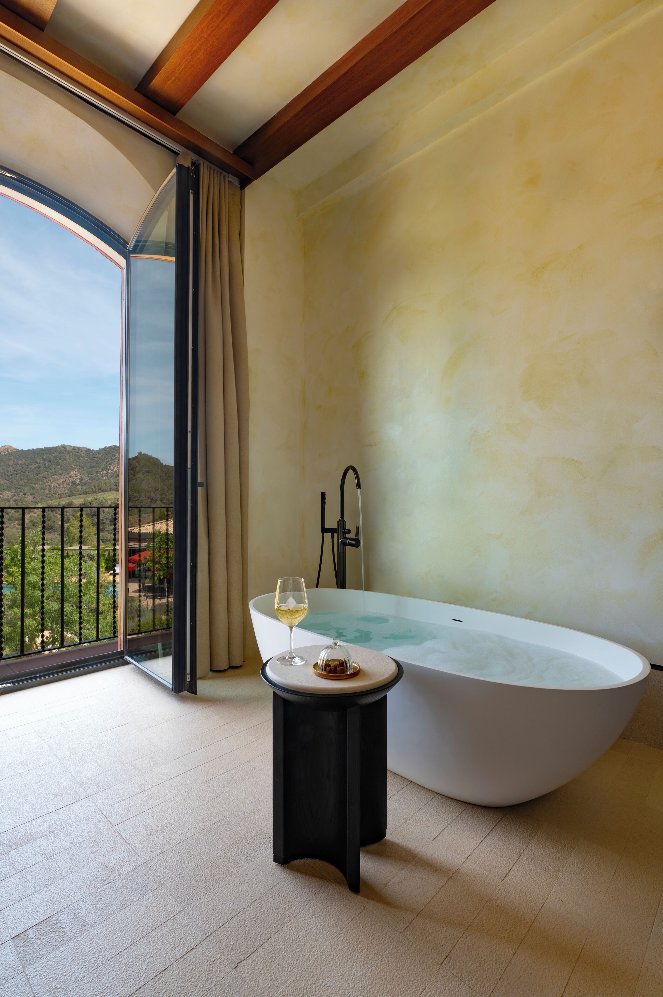 A modern bathroom with a freestanding bathtub, a small table with a glass of wine, and a view of a mountainous landscape through a large sliding door.