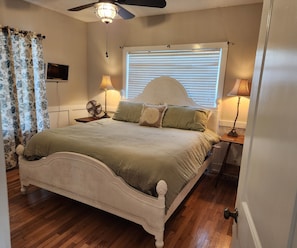 A bedroom with a white bed, two bedside lamps, a ceiling fan, a window with blinds, and a TV on the wall.