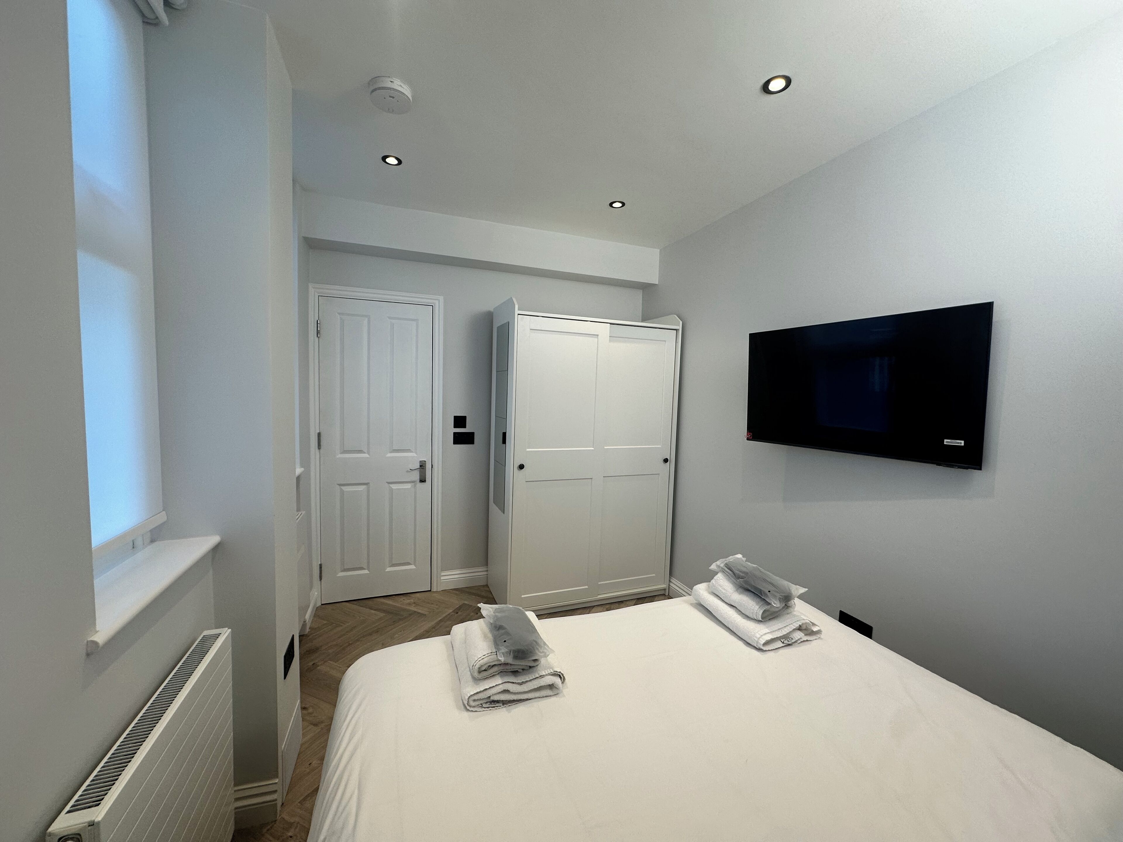 A hotel room with a bed, two white doors, a TV mounted on the wall, and a window.