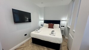 Una habitación de hotel moderna con una cama grande, un televisor de pantalla plana montado en la pared, mesitas de noche con lámparas y un piso de madera con patrón de espiga.