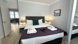 A modern hotel room with a large bed, bedside lamps, a flat-screen TV, and a view of the living area through an open door. - Chiltern Street Apartments - Superior E (London)