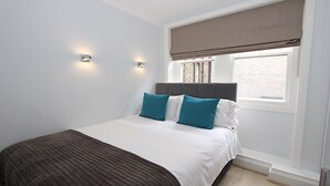 A bedroom with a bed, two wall-mounted lights, a window with a blind, and a view of a brick building outside. - Chiltern Street Apartments - Superior E (London)