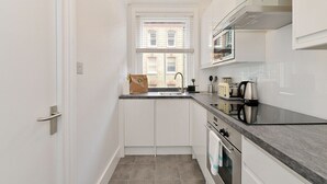 A compact kitchen with white cabinets, a built-in oven, a stainless steel range hood, and a window with blinds. - Chiltern Street Apartments - Superior E (London)
