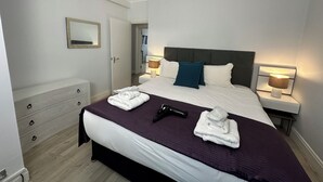 A neatly made bed with a purple bedspread, white pillows, and a dark headboard. There are two bedside lamps, a mirror on the wall, and a wooden dresser with drawers.