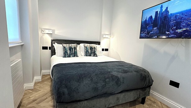 Quarto moderno com uma cama grande, piso de madeira e uma TV com paisagem urbana na parede.