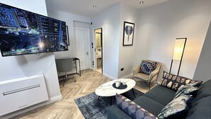 Uma sala de estar moderna com um sofá, uma mesa de centro, uma TV na parede e uma poltrona com almofadas estampadas.