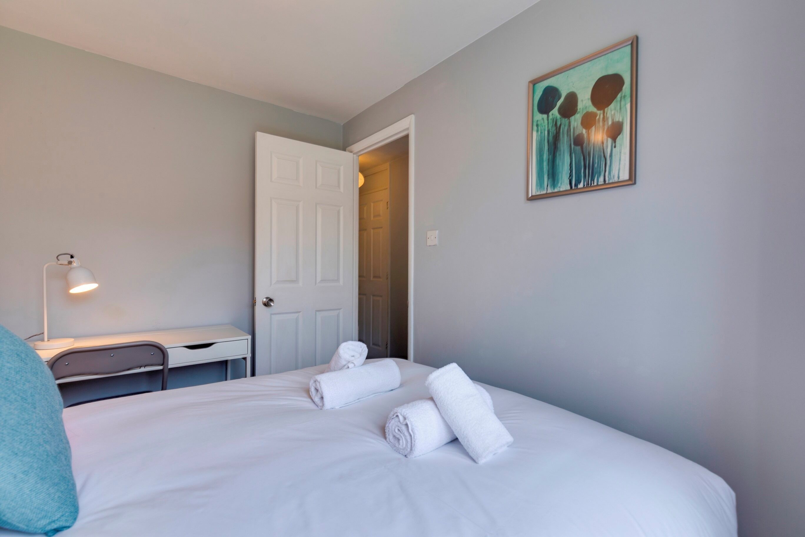 A hotel room with a bed, a desk, a chair, a lamp, and a painting on the wall.